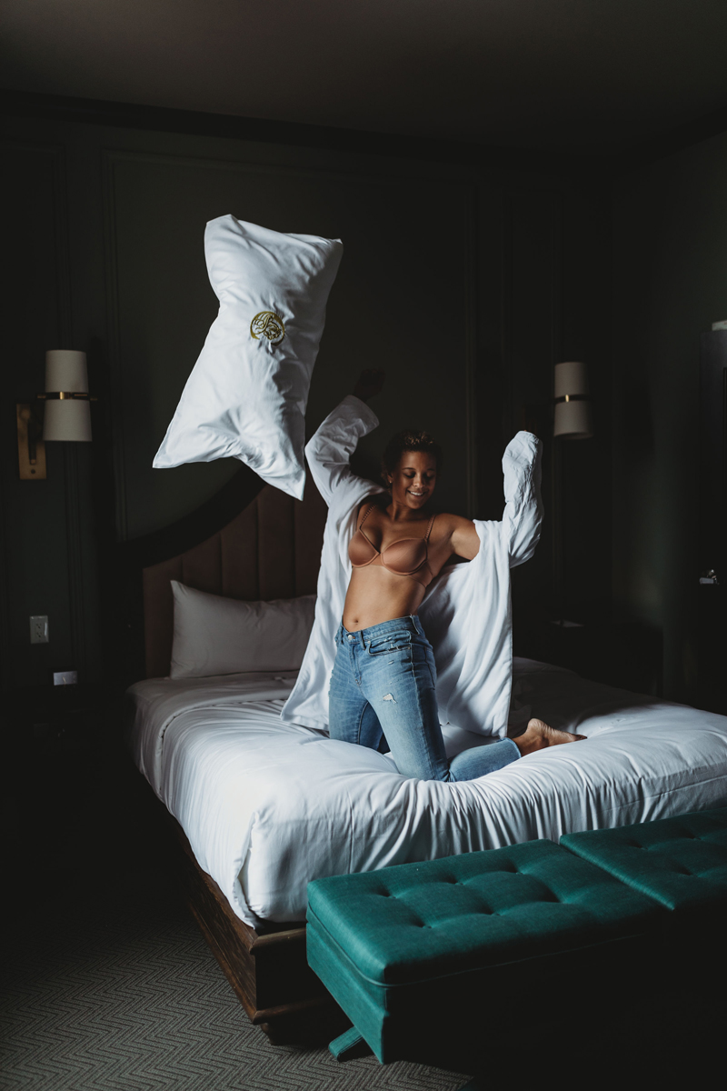 Atlanta Blogger-Influencer Photographer, on a bed, a woman with an open robe, bra, and jeans, tosses a pillow into the air