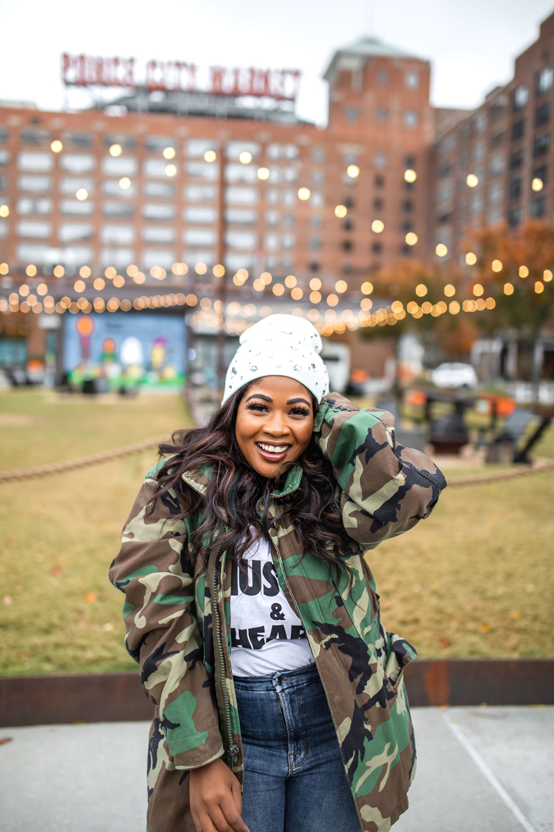 Atlanta Blogger-Influencer Photographer, a woman with a casual army style jacket smiles in a festive part of the city