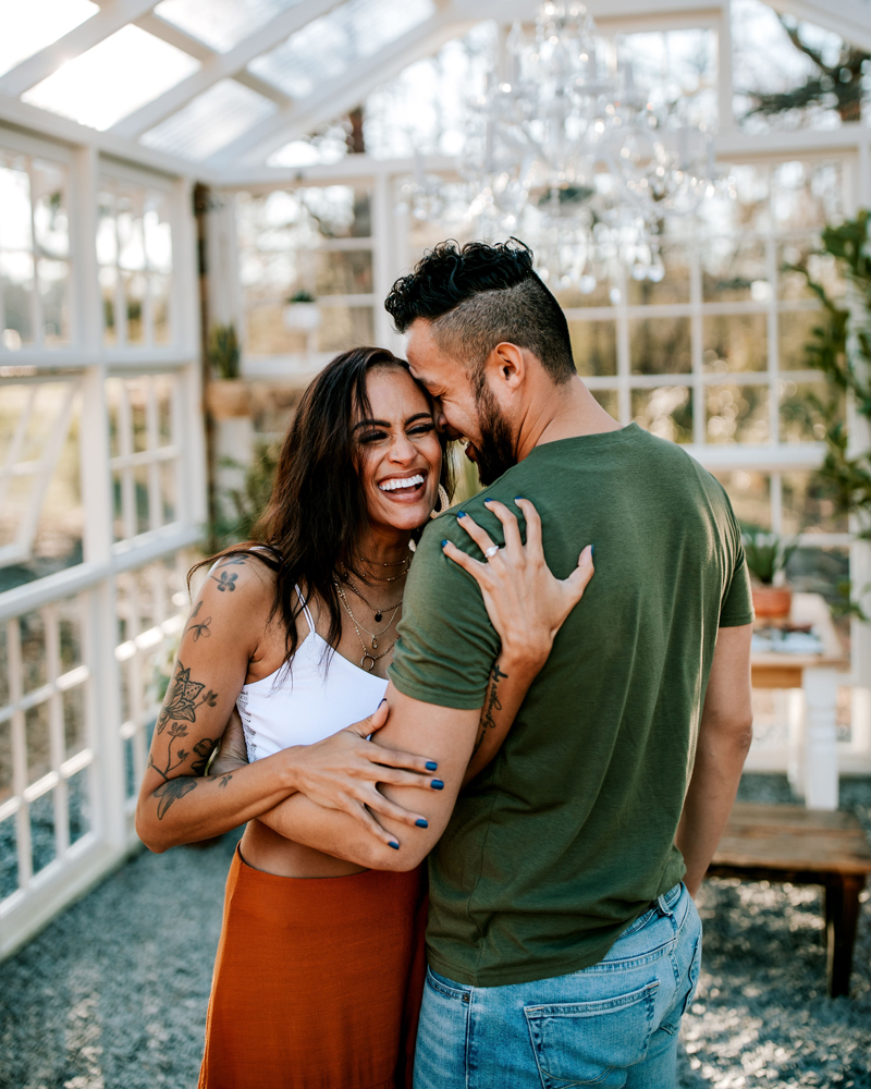 Atlanta Couples Photographer, couple embraces and smiles in greenhouse style patio. Woman has flower tattoos on arm.