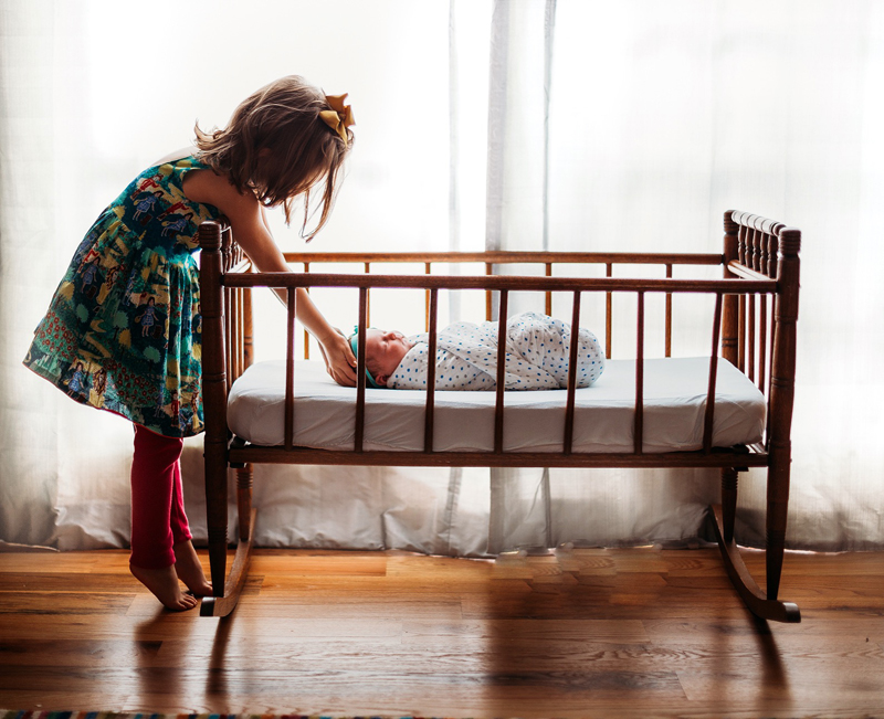 Atlanta Newborn Photographer, Toddler girl reaches into a wooden rocking crib to say hello to baby sister