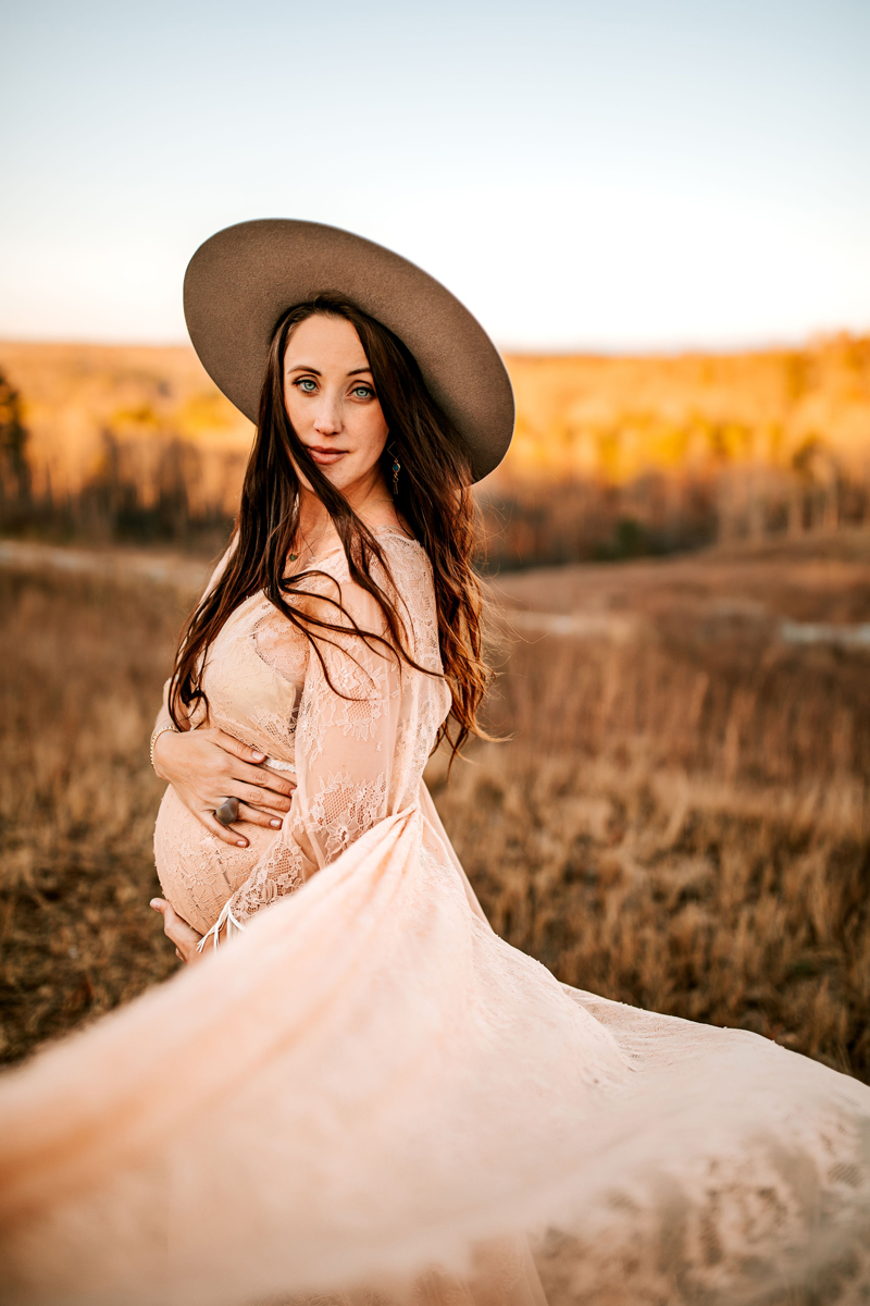 Atlanta Maternity Photographer, a pregnant woman holds her belly, she stands in a dry grassy field wearing a hat and a flowing dress