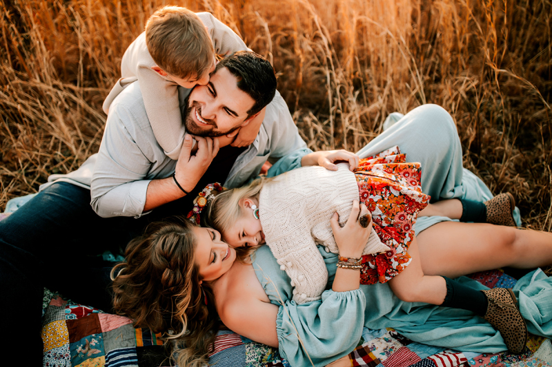 Atlanta Family Photographer, a boy hugs his dad and a girl climbs onto her mom as they all sit on a quilt in a grassy field