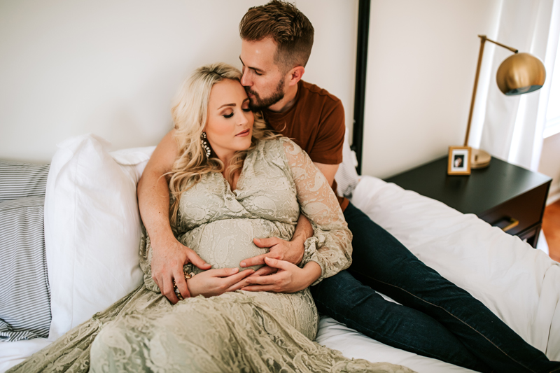 Atlanta Maternity Photographer, Man holds pregnant wife as they sit on their bed